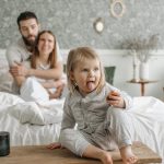 A cute toddler sitting on a bench in a cozy bedroom, with parents sitting on the bed.
