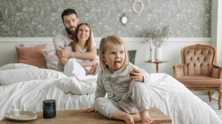 A cute toddler sitting on a bench in a cozy bedroom, with parents sitting on the bed.