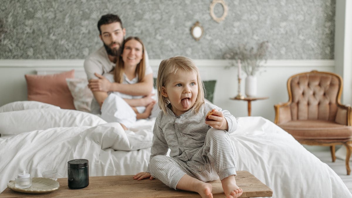 A cute toddler sitting on a bench in a cozy bedroom, with parents sitting on the bed.