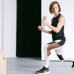A young woman performing lunges with dumbbells in a modern indoor gym setting.