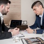 Two businessmen in suits discussing cryptocurrency trends using digital devices in an office setting.